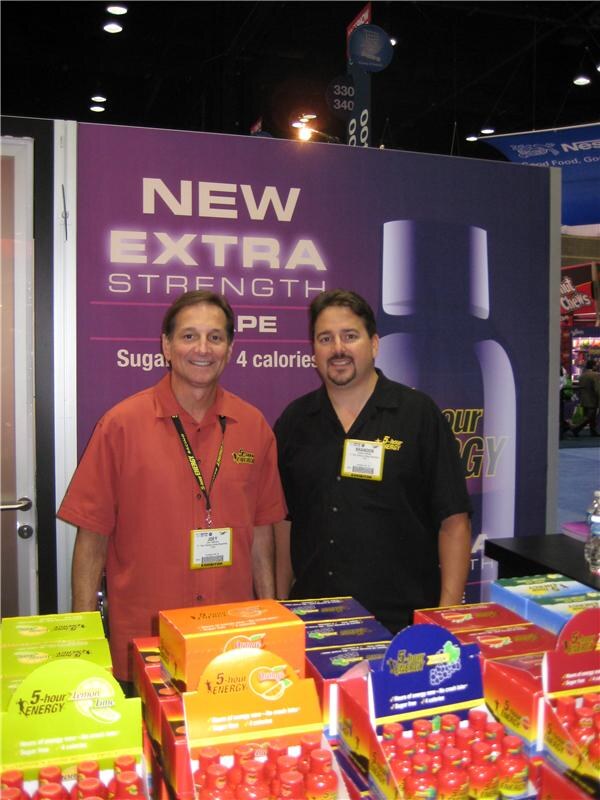 Joey, left, in an orange shirt and Brandon, right, in a black shirt stand in front of a 'New Extra Strength' backdrop above displays of 5-hour Energy shots.