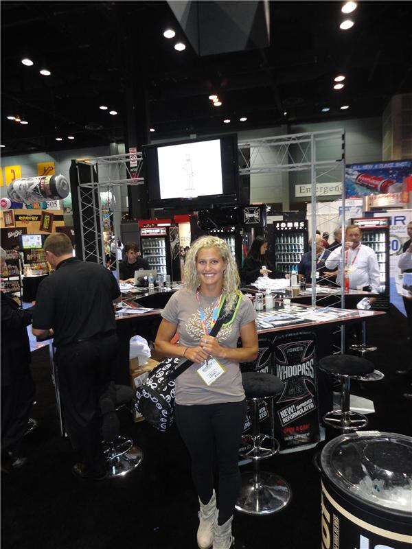 A smiling woman with curly blonde hair and a shoulder bag stands in front of the Jones Soda booth, where a Whoopass sign, bar stools, coolers and a video screen are visible.
