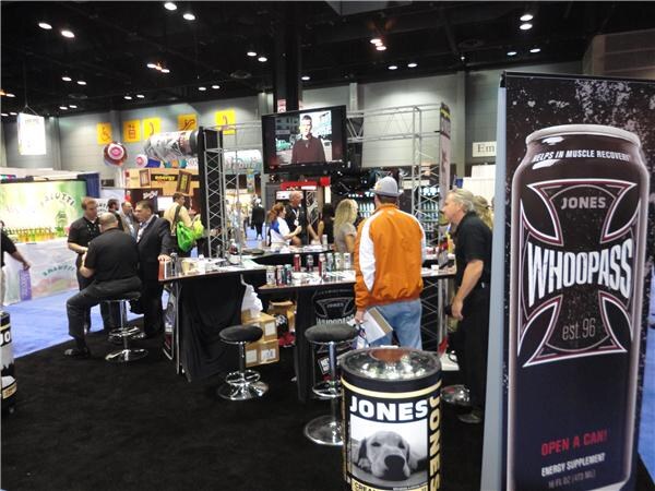 Visitors and staff gather around the Jones Soda booth, with a tall Whoopass can banner at right, a Jones barrel with a dog photo in front and a video screen above.