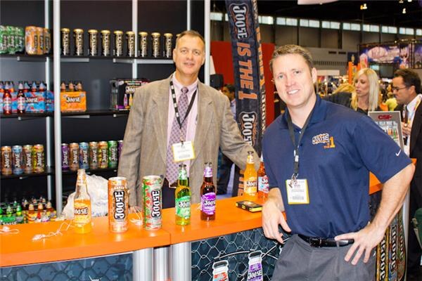Two men wearing badge lanyards stand at an orange counter holding Joose cans and bottles, with shelves of Joose products and a Joose banner behind them.
