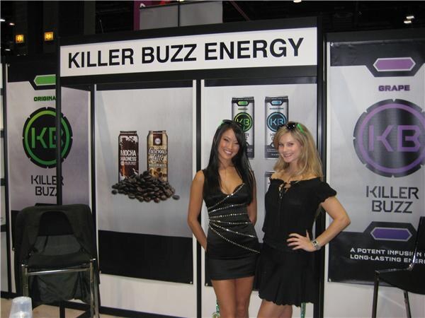Two women in black outfits stand in front of a Killer Buzz Energy sign and banners showing KB logos and cans of mocha, latte and grape flavors.