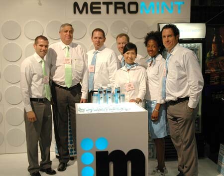 Six people in white and light-colored shirts stand around a white counter with water bottles, below a Metromint sign.