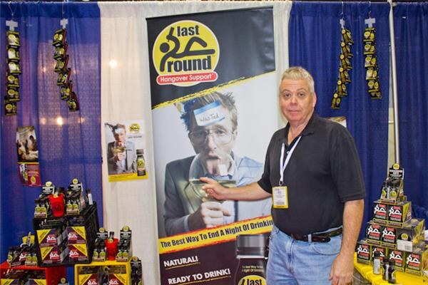 A man in a black polo with a lanyard badge stands beside a Last Round Hangover Support banner showing a man with a name tag on his forehead; boxed product is stacked on shelves and tables.