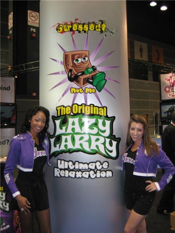 Two smiling women in purple jackets with Drank logos stand on either side of a tall column showing the Original Lazy Larry Ultimate Relaxation logo and a cartoon character.