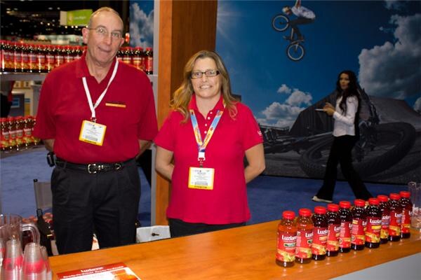 A man and a woman in red polo shirts and badge lanyards stand behind a wooden counter holding a row of Luzianne tea bottles, with bottles on shelves behind them.