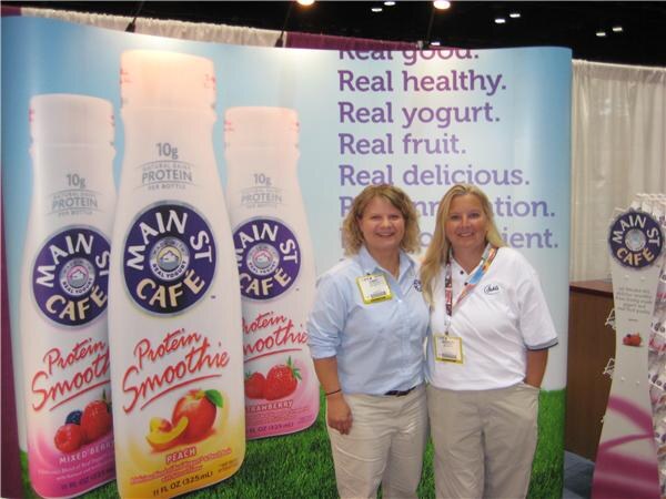 Two women in light shirts and khaki pants smile in front of a Main St. Cafe backdrop showing Protein Smoothie bottles in Mixed Berry, Peach and Strawberry.