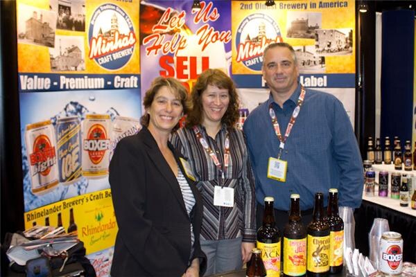 Two women and a man in conference badges stand together at the Minhas Craft Brewery booth, with Boxer cans on the banner and Rhinelander craft beer bottles on the table in front.