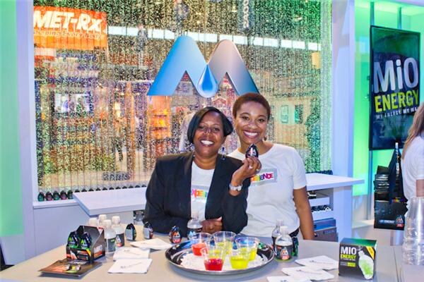 Two smiling women sit behind a counter with small cups of colorful drinks and MiO Energy boxes, with a MiO logo, MET-Rx sign and MiO Energy banner behind them.