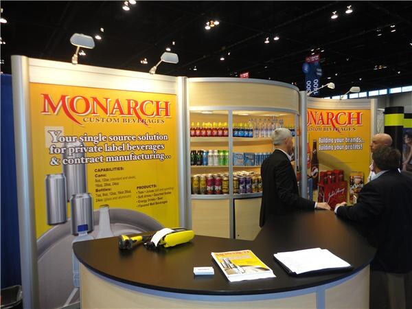 Two men in dark suits talk at a curved counter at the Monarch Custom Beverages booth, which has yellow banners about private label beverages and a lit shelf of bottled and canned drinks.