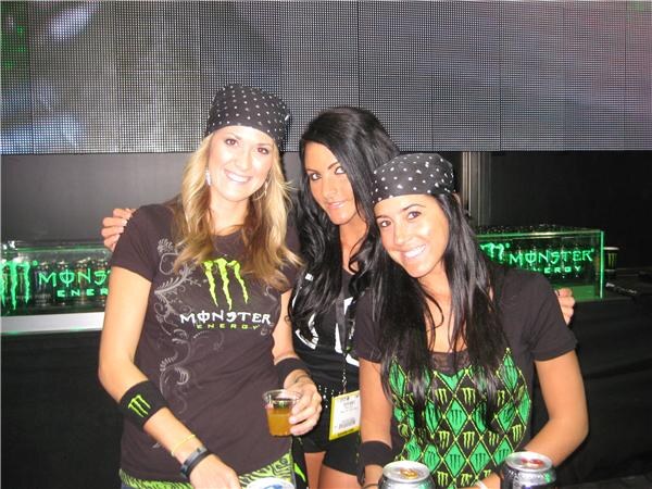 Three smiling women in Monster Energy clothing and black bandanas stand together behind a counter with Monster cans, with green-lit Monster Energy displays behind them.