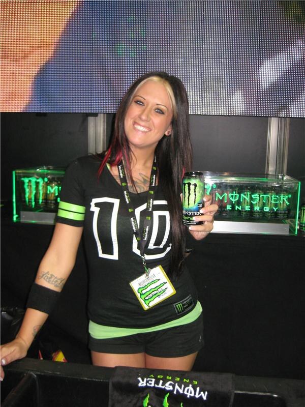 A smiling woman with dark hair and a lanyard badge, wearing a black Monster Energy shirt, holds a green Monster can in front of lit Monster Energy display cases.