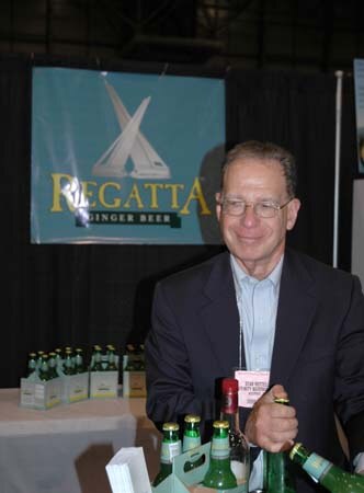 A man in glasses and a dark jacket stands behind a table holding green bottles, with a Regatta Ginger Beer banner behind him.