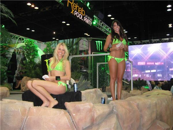 Two women in green bikinis, one seated and one standing, on faux rock formations at a Rehab Las Vegas Pool Party display with a Monster Energy logo, plants and a video screen behind them.