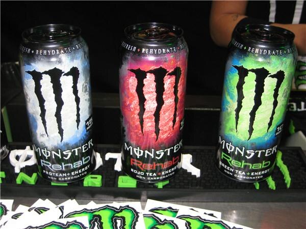 Three Monster Rehab cans stand on a black bar mat: Protean plus Energy in blue-white, Rojo Tea plus Energy in red, and Green Tea plus Energy in green.