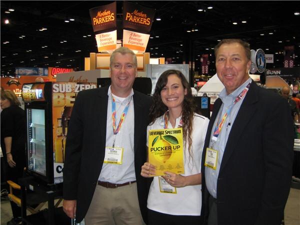 Two men and a woman in a Mother Parkers booth; the woman holds a Beverage Spectrum magazine with a lemon on the cover. A Bunn sign is visible behind.