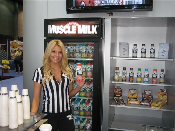 A smiling woman with long blonde hair in a striped referee-style shirt holds a Muscle Milk bottle beside a lit Muscle Milk cooler. A shelf of bottles and bars stands to the right.