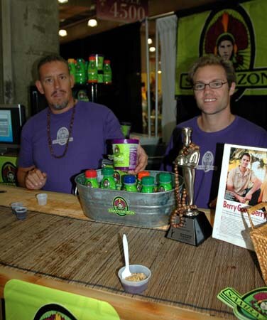 Two men in purple shirts stand behind a counter with a tub of green-capped Sambazon bottles, a small trophy and a magazine page, with a Sambazon banner behind.