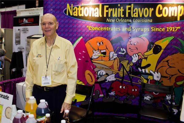 A smiling bald man in a yellow shirt and lanyard stands at a booth with bottles of syrup on the table. Behind him is a purple cartoon fruit banner reading National Fruit Flavor Company, New Orleans, Louisiana.