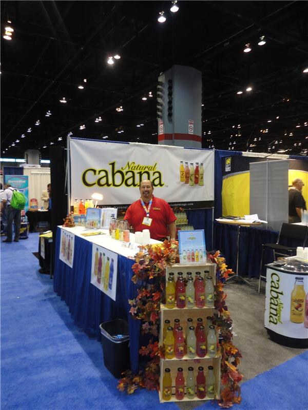 A man in a red shirt stands behind a counter under a Natural Cabana banner, with crates of bottled lemonade decorated with autumn leaves in front of the booth.
