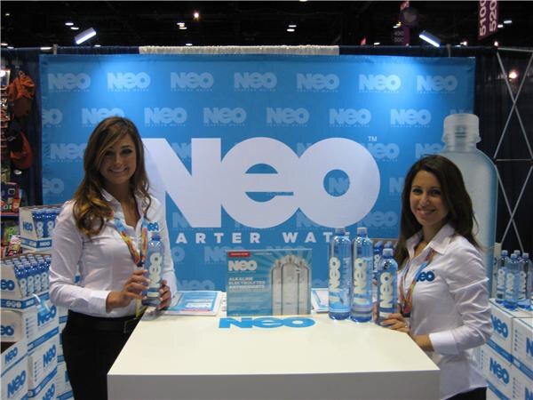 Two smiling women in white shirts stand behind a white counter displaying NEO water bottles, in front of a large blue NEO backdrop. Cases of NEO water are stacked at either side.