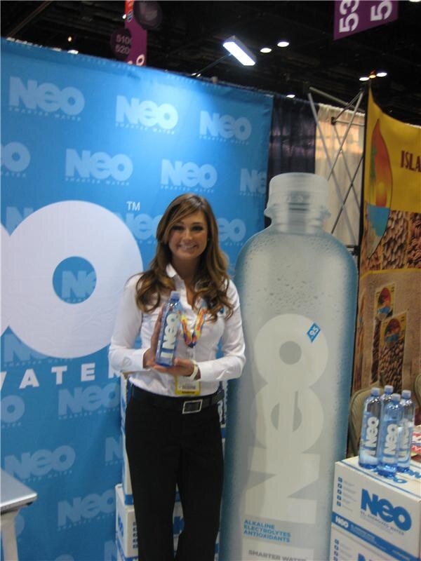 A smiling woman in a white shirt and lanyard holds a bottle of NEO water beside a giant NEO bottle display and a blue NEO backdrop. Cases of NEO water sit nearby.