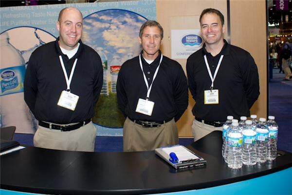 Three smiling men in black polo shirts and badge lanyards stand behind a curved black counter at the Nestlé Waters booth. Bottles of water and a clipboard sit on the counter, and a Nestlé Pure Life graphic is behind them.