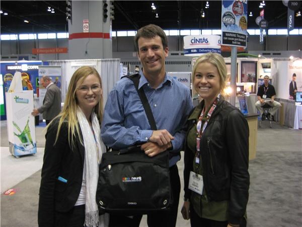 A man in a blue checked shirt holding a black Neuro-branded messenger bag poses between two smiling women on a trade-show floor. A Cintas sign hangs in the background.