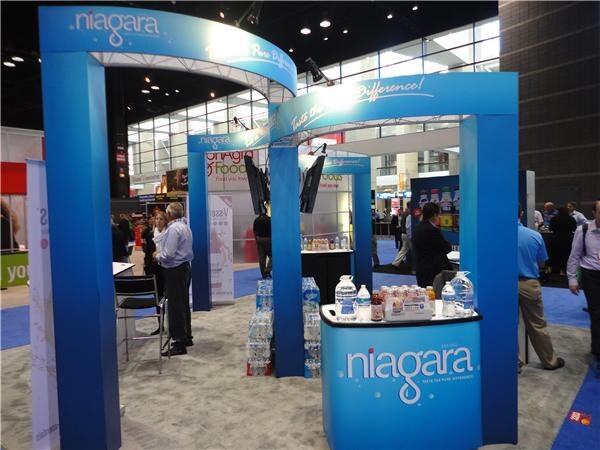 A blue Niagara booth with curved arches and a counter displaying bottled water. Stacked cases of water stand nearby, and several attendees walk through the aisle of the show floor.