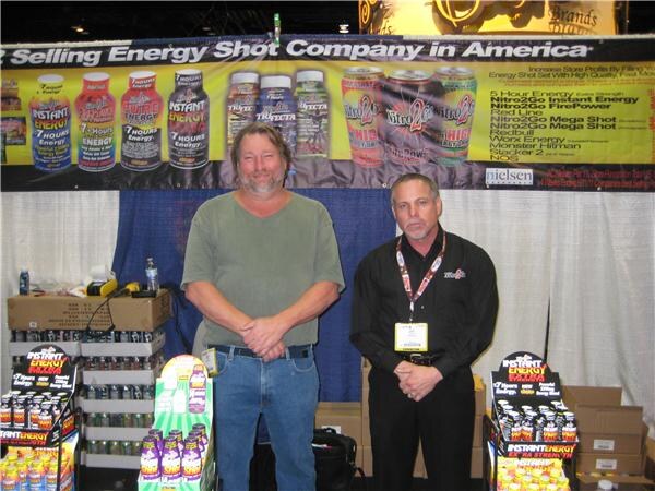 Two men stand in front of a banner showing Instant Energy and Nitro2Go energy shot products, with Instant Energy product displays and boxes on either side of them.