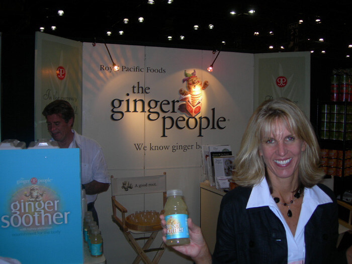 A smiling blonde woman holds a bottle of Ginger Soother in front of The Ginger People and Royal Pacific Foods sign. A man stands behind the counter at left.