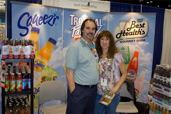 A man and a woman wearing lanyards pose together in the North Shore Bottling booth, in front of banners for Squeez'r, Tropical Fantasy and Best Health's Gourmet Soda, with bottles on shelves.