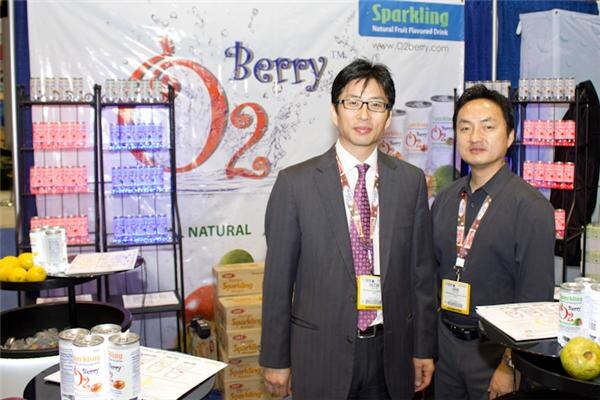 Two men in suits and badges stand in front of an O2 Berry sparkling fruit drink banner, with cans of O2 Berry on tables in front and shelves of drinks behind them.