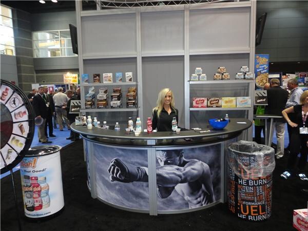 A woman with long blonde hair stands behind a curved counter at the Oh Yeah! booth, with product boxes on shelves, bottles on the counter, a prize wheel at left and a boxer graphic on the front.