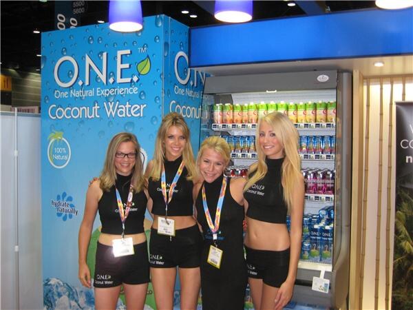 Four women in matching black O.N.E. Coconut Water outfits and lanyards stand together in front of a blue O.N.E. banner and a refrigerated display case filled with bottled drinks.