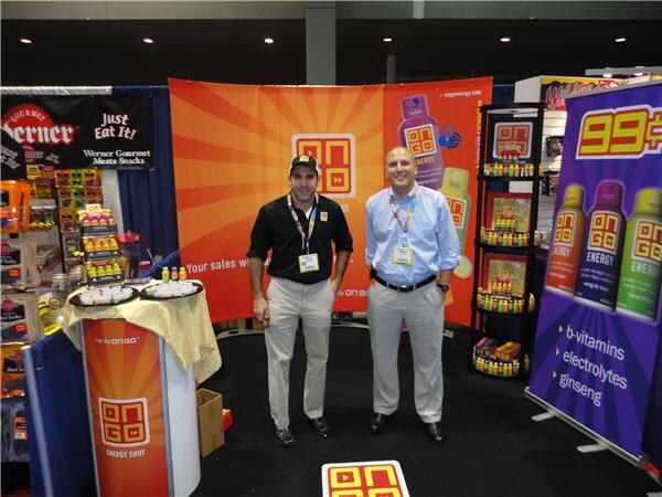 Two men with lanyards stand in an OnGo energy shot booth with orange banners and shelves of shots. A Werner Gourmet Meats Snacks booth is visible to the left.