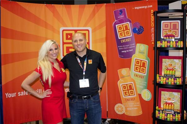A blonde woman in a red dress and a man in a black shirt with a lanyard stand together in front of orange OnGo Energy banners and shelves of energy shots.