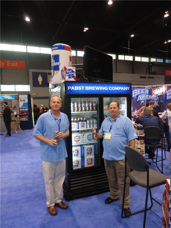 Two men in light blue shirts stand beside a Pabst Brewing Company cooler stocked with Pabst Blue Ribbon, Old Milwaukee and Lone Star, with a large inflatable can above it.