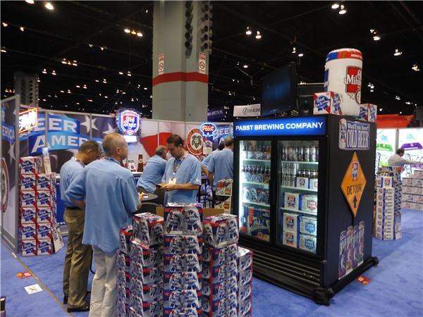 Men in light blue shirts stand near stacked cases of Pabst Blue Ribbon and a Pabst Brewing Company cooler. Old Style and Oly signs appear in the busy booth.