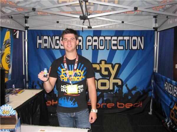 A smiling young man in a Party Armor t-shirt and lanyard holds up a small bottle in a tent booth with a blue Hangover Protection banner behind him.