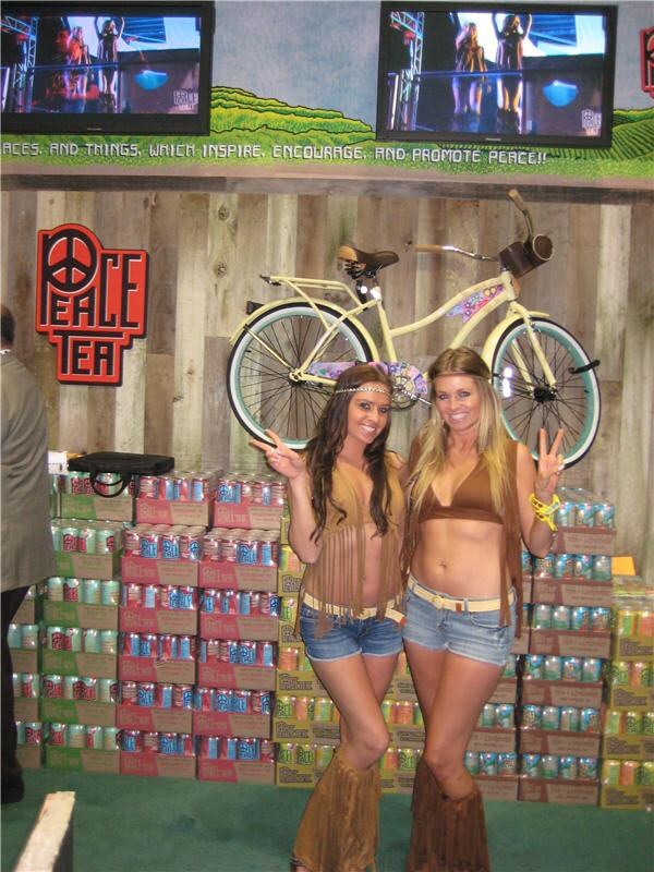 Two women in fringed western-style outfits flash peace signs in front of stacked Peace Tea cases, a Peace Tea sign, and a cream bicycle mounted on a wood wall.