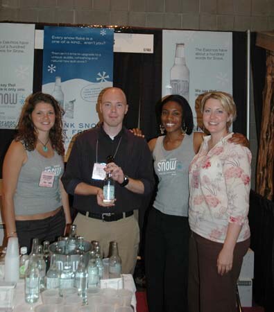Three women and a bald man holding a bottle stand together at a Snow booth with banners behind them and bottles and cups on the table in front.