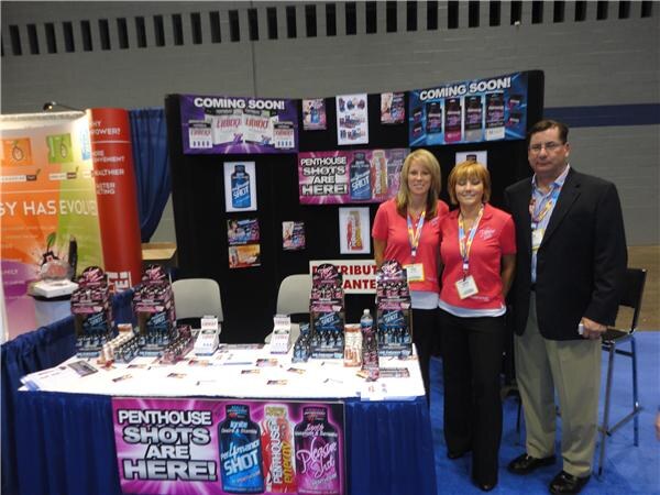 Two women in pink shirts and a man in a dark blazer stand at a booth table with Penthouse Shots displays and signs reading Penthouse Shots Are Here and Coming Soon.