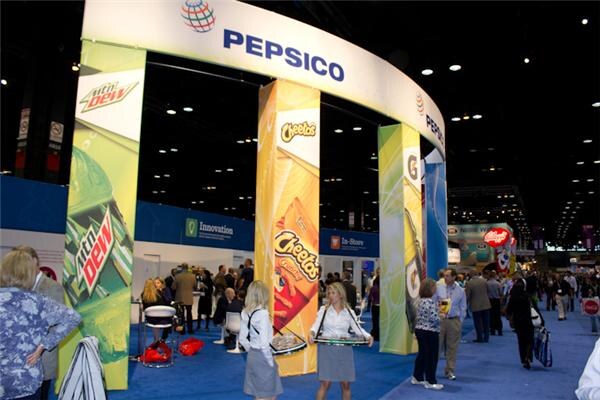 Large curved PepsiCo booth entrance with Mountain Dew and Cheetos banners on the pillars, with attendees and two women holding trays in the foreground on the trade show floor.