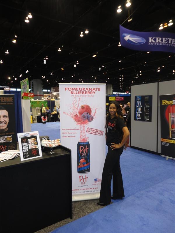 A woman in a black shirt and pants stands beside a Pit Bull Energy Drink Pomegranate Blueberry banner in a booth, with a Kretek International sign hanging overhead.