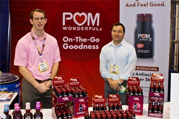 Two men wearing badges stand behind a table of POM Wonderful bottles and POM Shot X displays, in front of a red POM Wonderful sign reading On-The-Go Goodness.