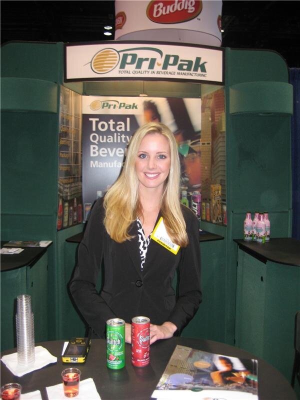 A smiling woman with long blonde hair stands at the Pri-Pak booth holding her hands near two cans of Switch on a table, with a Buddig sign above the booth.