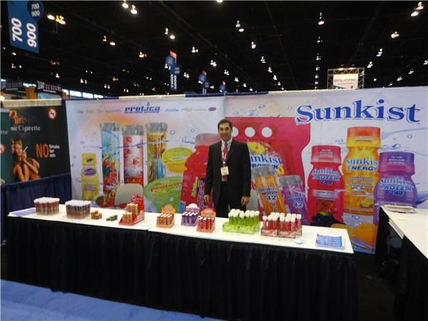 A man in a dark suit stands behind a table of small bottles in front of a large backdrop showing Protica and Sunkist protein and energy drinks.