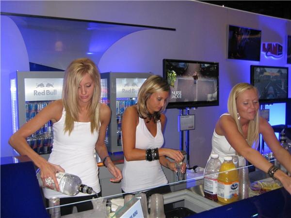 Three women in white tops work behind a bar, one pouring from a bottle, with Red Bull coolers and video screens behind them under blue lighting.