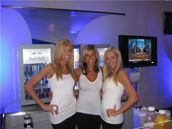Three smiling women in white tops pose together in front of a lit Red Bull cooler and a television screen in a blue-lit booth.