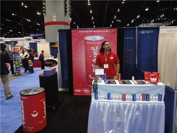 A man in a red shirt stands at a draped table with Cocaine energy drink cans, beside a tall red Cocaine can banner and a Galaxy Life sign in the background.